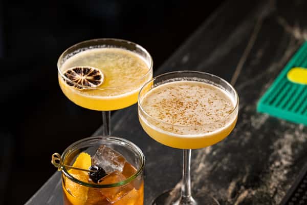 Trio of craft cocktails on a dark bar counter, including two coupe glasses with citrus-forward drinks—one garnished with a lime wheel and the other with spice—alongside a classic Old Fashioned with orange peel and cherry.