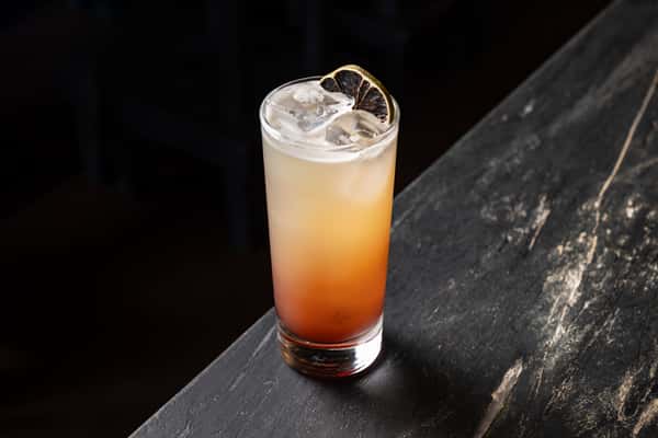 Alternate view of a vibrant mezcal cocktail with ice and charred lime wheel, served in a highball glass with a warm gradient tone on a dark marble bar.