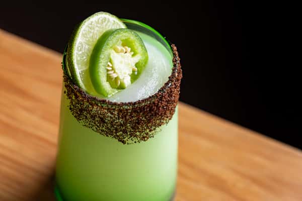 Frozen margarita in a green-rimmed glass, garnished with jalapeño and lime slices and a chili-lime rim, served on a wooden bar