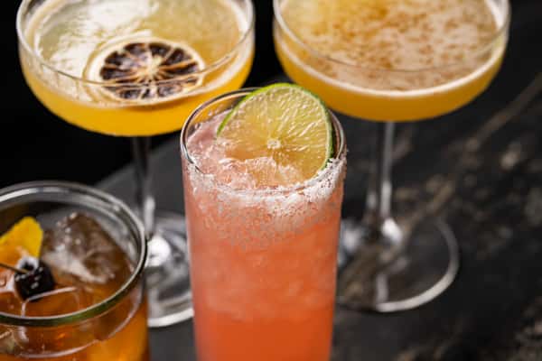 Centerpiece pink mezcal cocktail with salt rim and lime wedge, surrounded by citrus-forward drinks in coupe and lowball glasses on a stone bar.