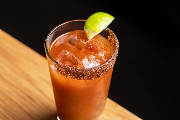 Close-up of a classic michelada cocktail in a tall glass with chili-lime rim, ice cubes, and fresh lime garnish, served on a wooden bar at La Hol