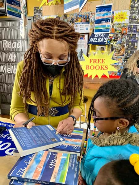 Woman signing books for a child at Melba's community bookstore with "Free Books" display and colorful reading posters in the background