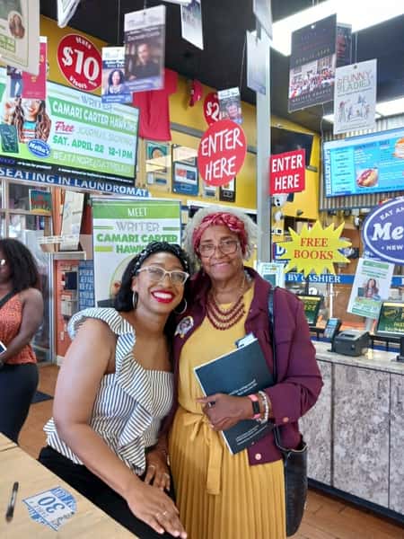 two women posing together at Melba's restaurant bookstore during author event with "Enter Here" and "Free Books" signs