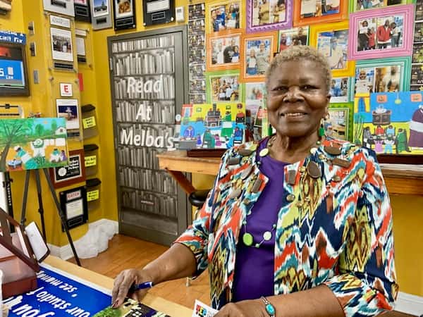 author smiling at book signing table inside Melba's restaurant bookstore with colorful "Read at Melba's" wall and community artwork