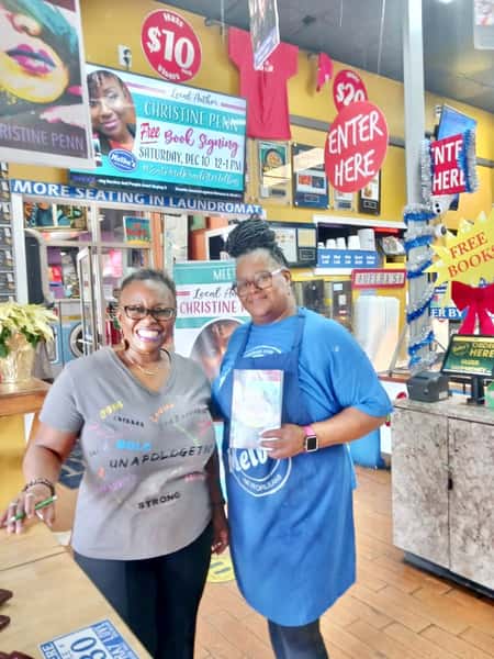 two women posing with book at Melba's restaurant bookstore during local author signing event with "Free Books" signs