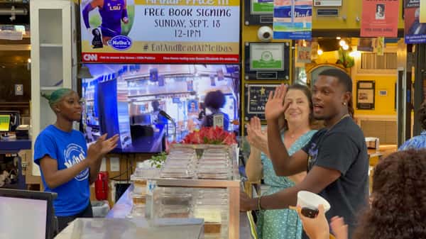 Melba's staff and guests clapping near takeout counter during live book event with Southern food containers and event signage