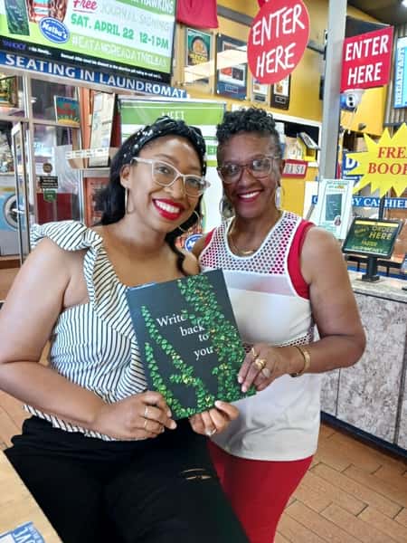 two women smiling and holding a book inside Melba's restaurant with "Free Books" signs and vibrant community atmosphere
