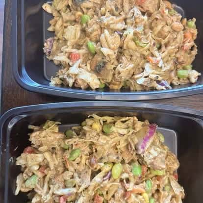 Shredded chicken salad with cabbage, edamame, and red onions in a creamy dressing, packed in two black meal prep containers.