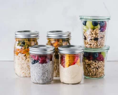 Mason jars filled with rice, chia pudding, hummus, fruits, vegetables, and granola stacked neatly on a white surface.