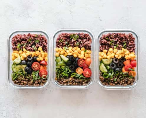 Colorful vegan meal prep bowls with black rice, corn, cherry tomatoes, cucumber, olives, and green onion in glass containers.