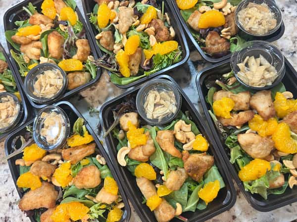 Meal prep containers with leafy green salad, mandarin oranges, cashews, crispy chicken pieces, and sealed cups of crunchy toppings.