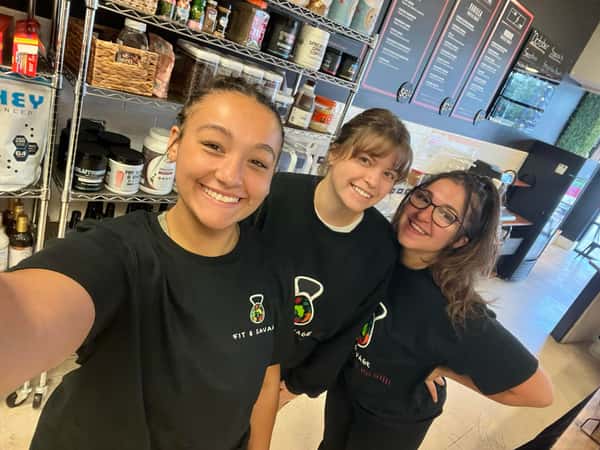 Three smiling women wearing black "Fit & Savage" shirts posing inside a supplement and meal prep shop.