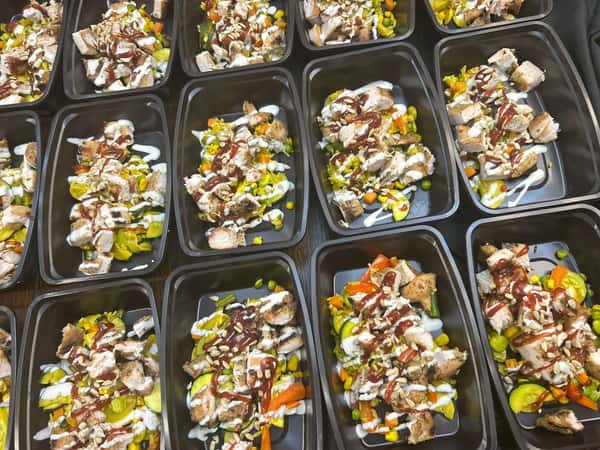 Meal prep containers with grilled chicken, mixed vegetables, pickles, ranch and BBQ drizzle, and sunflower seeds.