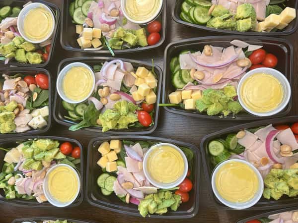 Meal prep trays filled with turkey, avocado, cherry tomatoes, cucumber, cheese cubes, red onion, and almonds with dressing cups.
