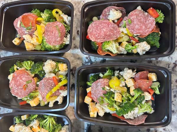 Meal prep trays with broccoli, cauliflower, salami, pepperoni, cheddar cubes, and sliced peppers on a marbled counter.
