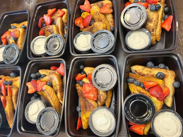French toast meal prep containers with sliced strawberries, blueberries, and individual syrup and cream sauce portions.