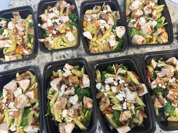 Penne pasta meal prep bowls with grilled chicken, wilted spinach, sun-dried tomatoes, and crumbled feta in black containers.