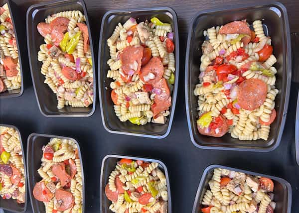 Meal prep containers filled with rotini pasta salad, pepperoni slices, diced chicken, red onions, peppers, and cherry tomatoes.