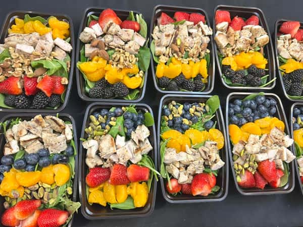 Grilled chicken salad bowls with spinach, mandarin oranges, berries, strawberries, and nuts, prepped in black meal containers.