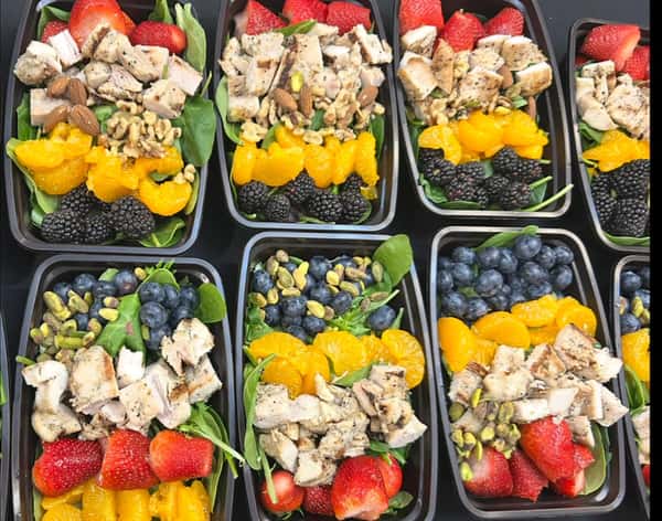 Meal prep containers filled with spinach, grilled chicken, berries, mandarin oranges, and mixed nuts arranged on a black surface.