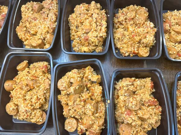 Meal prep containers filled with rice, sliced sausage, tomatoes, and seasonings, resembling a jambalaya-style dish.