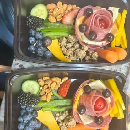 Colorful lunch trays with salami rose, blueberries, cucumber, nuts, cheese slices, bell pepper sticks, and granola clusters.