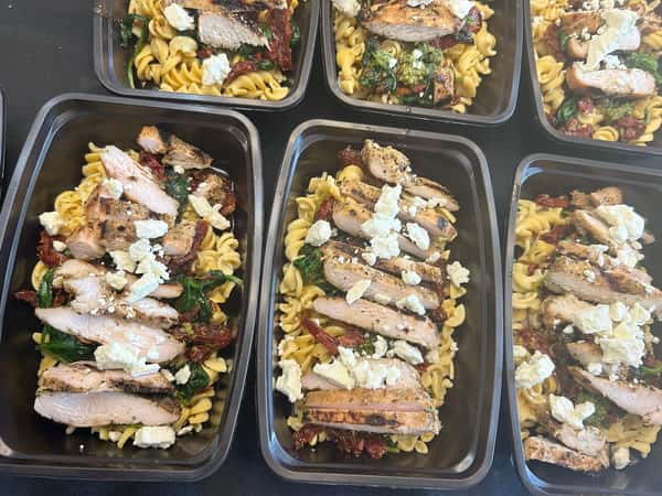 Grilled chicken with sun-dried tomatoes, spinach, rotini pasta, and crumbled feta cheese in meal prep containers.