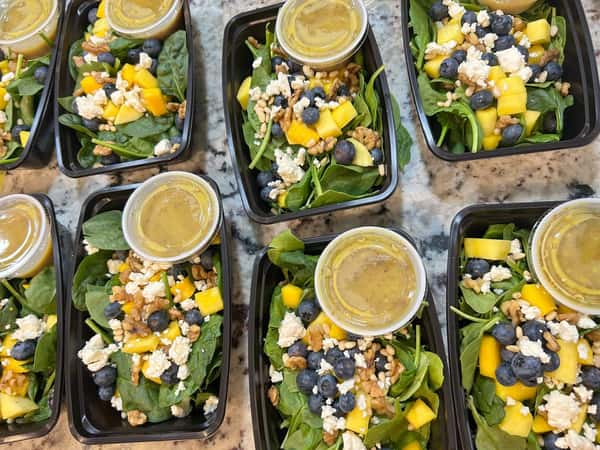 Fresh spinach salad meal prep with blueberries, mango chunks, feta, walnuts, and dressing cups in black containers.