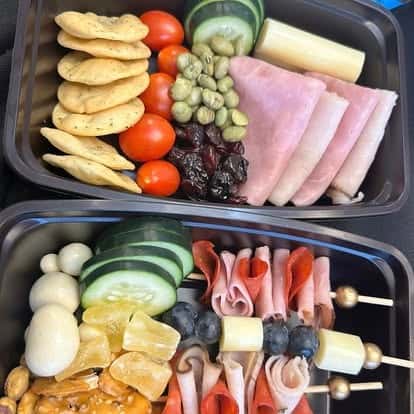 Assorted lunch trays with crackers, ham slices, cucumber, tomatoes, dried fruit, cheese cubes, and charcuterie skewers.
