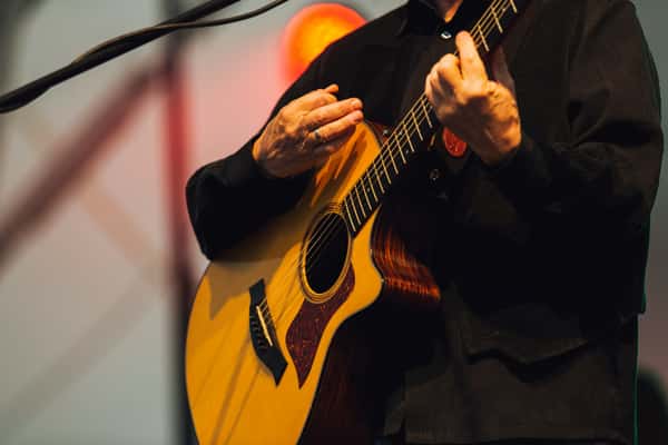 Close-up of a person playing an acoustic guitar on stage, wearing a black shirt with stage lighting in the background.