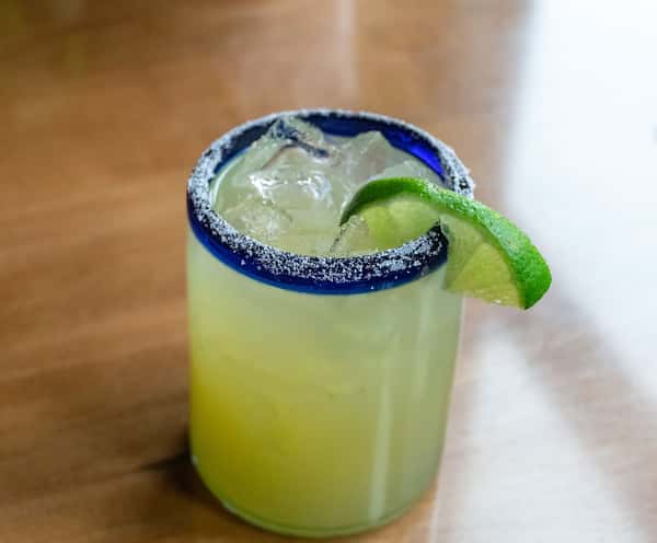 Salt-rimmed margarita with lime wedge and ice in a blue-rim glass, served on a wooden table.
