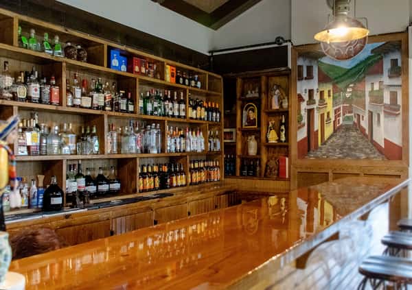 Rustic bar interior with wooden shelves stocked with liquor bottles, polished wood counter, and colorful mural of a Mexican village street.