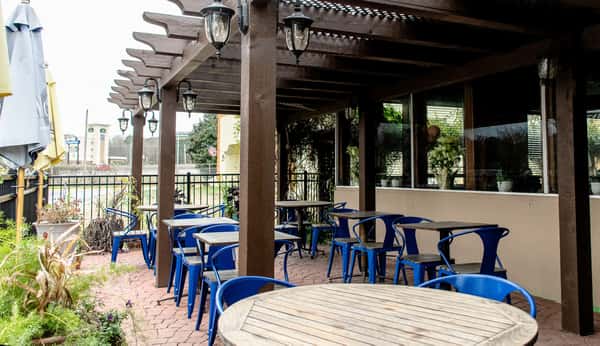 Covered outdoor restaurant patio with wooden pergola, lantern lights, brick flooring, and blue metal chairs around small dining tables.