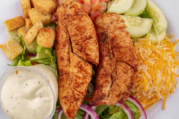 Grilled chicken salad with shredded cheese, cucumbers, croutons, red onions, lettuce, and a side of ranch dressing.