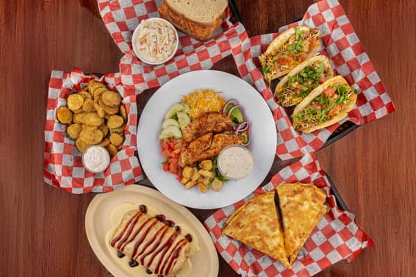 Assorted restaurant dishes including fried pickles, chicken salad, tacos, quesadilla, sandwich with coleslaw, and a smothered burrito.