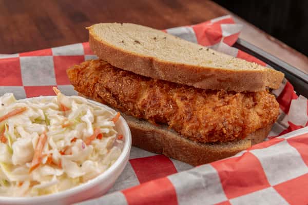 Crispy fried pork sandwich on rye bread served with a side of creamy coleslaw on red-and-white checkered paper.