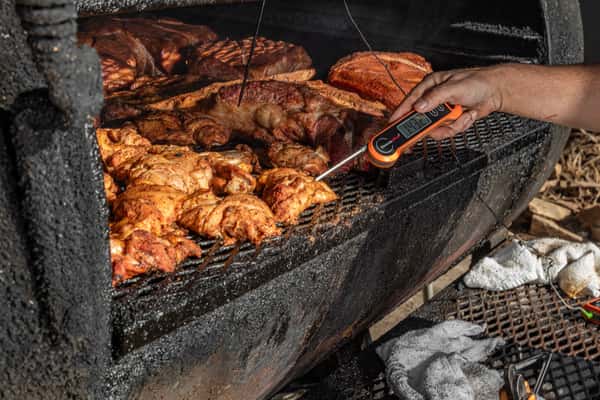 Barbecue pitmaster checking the temperature of smoked chicken with a digital thermometer inside a smoker filled with meats.