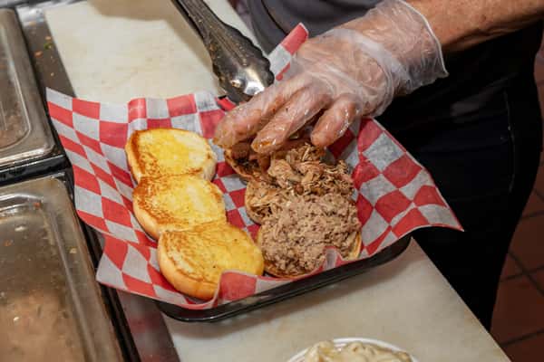 Gloved hands preparing a pulled pork sandwich on toasted buns, with shredded barbecue pork piled onto the bread in a basket.