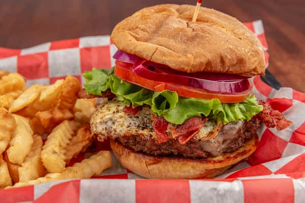 Cheeseburger topped with bacon, fried egg, lettuce, tomato, and onion, served with crinkle-cut fries on checkered paper.