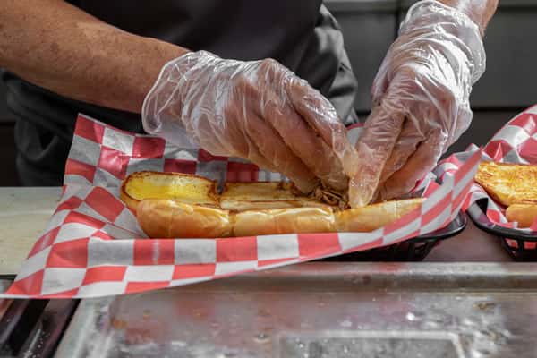 Person wearing gloves assembling a pulled pork sandwich on a toasted bun lined with red-and-white checkered paper.