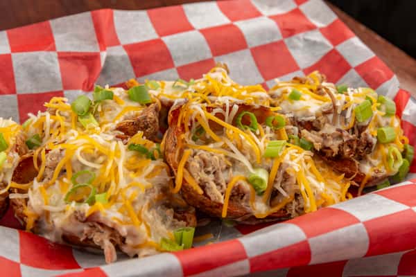 Loaded potato skins topped with shredded cheese, pulled pork, and sliced green onions on red-and-white checkered paper.