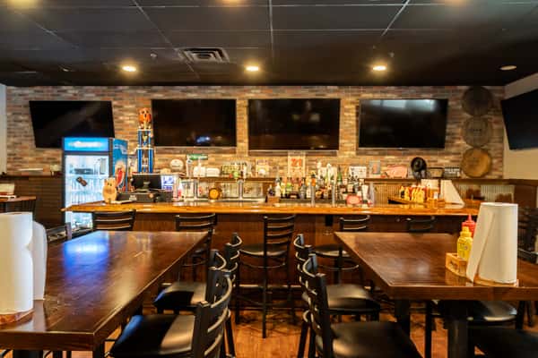 Bar and dining area with wooden tables, condiment caddies, multiple TVs on a brick wall, and a fully stocked bar with stools.