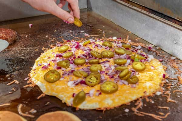 Cheese crisp topped with chopped red onions and jalapeño slices being prepared on a flat-top grill.
