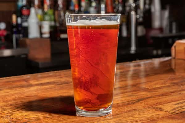 Pint glass of amber beer served on a wooden bar counter with bottles in the blurred background.