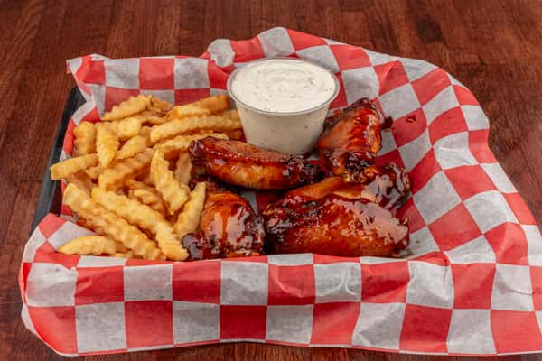 Basket of glazed barbecue chicken wings with crinkle-cut fries and ranch dipping sauce on red-and-white checkered paper.