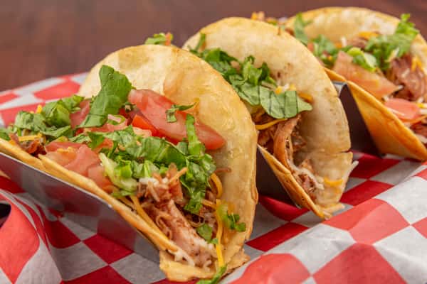 Three crispy tacos filled with shredded meat, lettuce, tomatoes, and cheese served on red-and-white checkered paper.