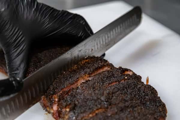 Close-up of black-gloved hands slicing a smoked beef brisket with a peppery bark—ideal for showcasing Texas BBQ technique or behind-the-scenes kitchen prep.