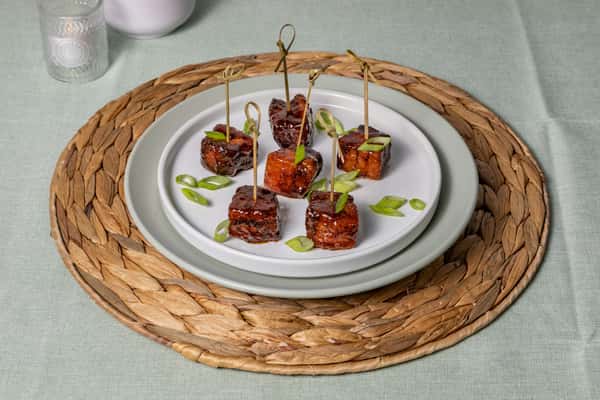 Barbecue-glazed pork belly bites skewered and topped with scallions, presented on a double-stacked white plate with a woven placemat—ideal for Southern small bites or modern BBQ starters.