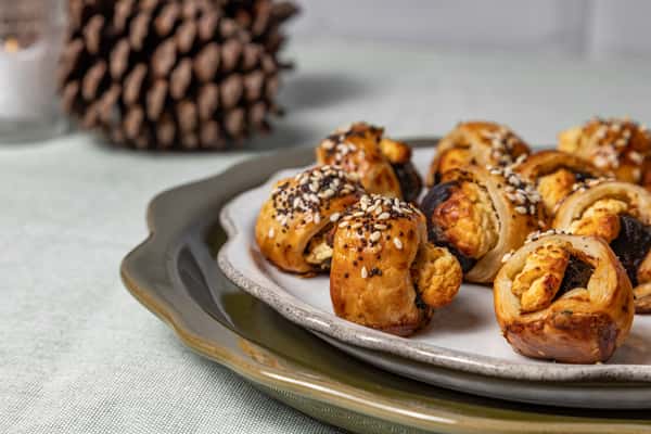 Close-up view of flaky puff pastry rolls with a golden finish, sprinkled with sesame seeds and served on layered ceramic plates—perfect for holiday platters or bite-sized snacks.