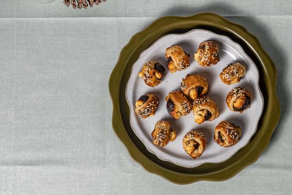 Golden-brown puff pastry bites filled with a savory filling and topped with sesame seeds, neatly arranged on a scalloped ceramic plate—ideal for catering menus or holiday party appetizers.